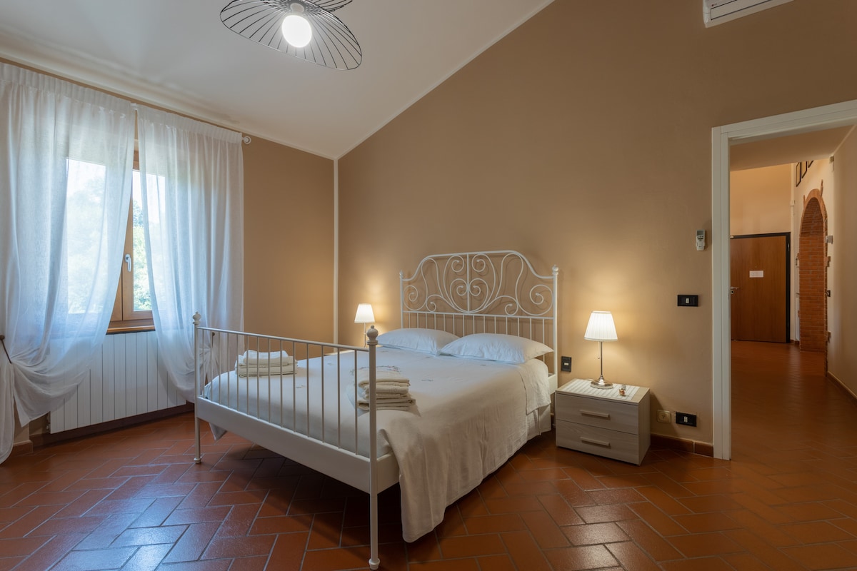 A spacious bedroom is highlighted by a metal bed frame and soft, neutral-toned walls. Two nightstands with lamps are positioned on either side of the bed. Natural light filters through the sheer curtains covering a large window, illuminating the terracotta tile floor.