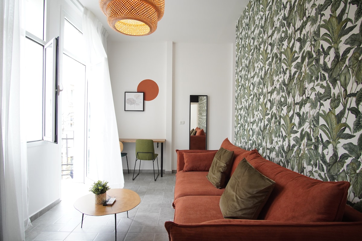A spacious living area features a plush orange sofa adorned with green cushions. A round coffee table sits in front, and a work desk with green chairs is visible. Natural light floods in through large windows, where light, flowing curtains add softness. A patterned accent wall enhances the ambiance.