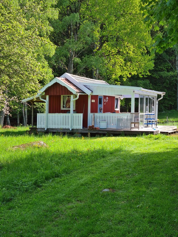 A small red cottage is surrounded by lush greenery, with a covered porch that features white railings. The structure includes large windows that provide ample natural light, and a pathway leads to the entrance, enhancing the cottage's inviting appearance.