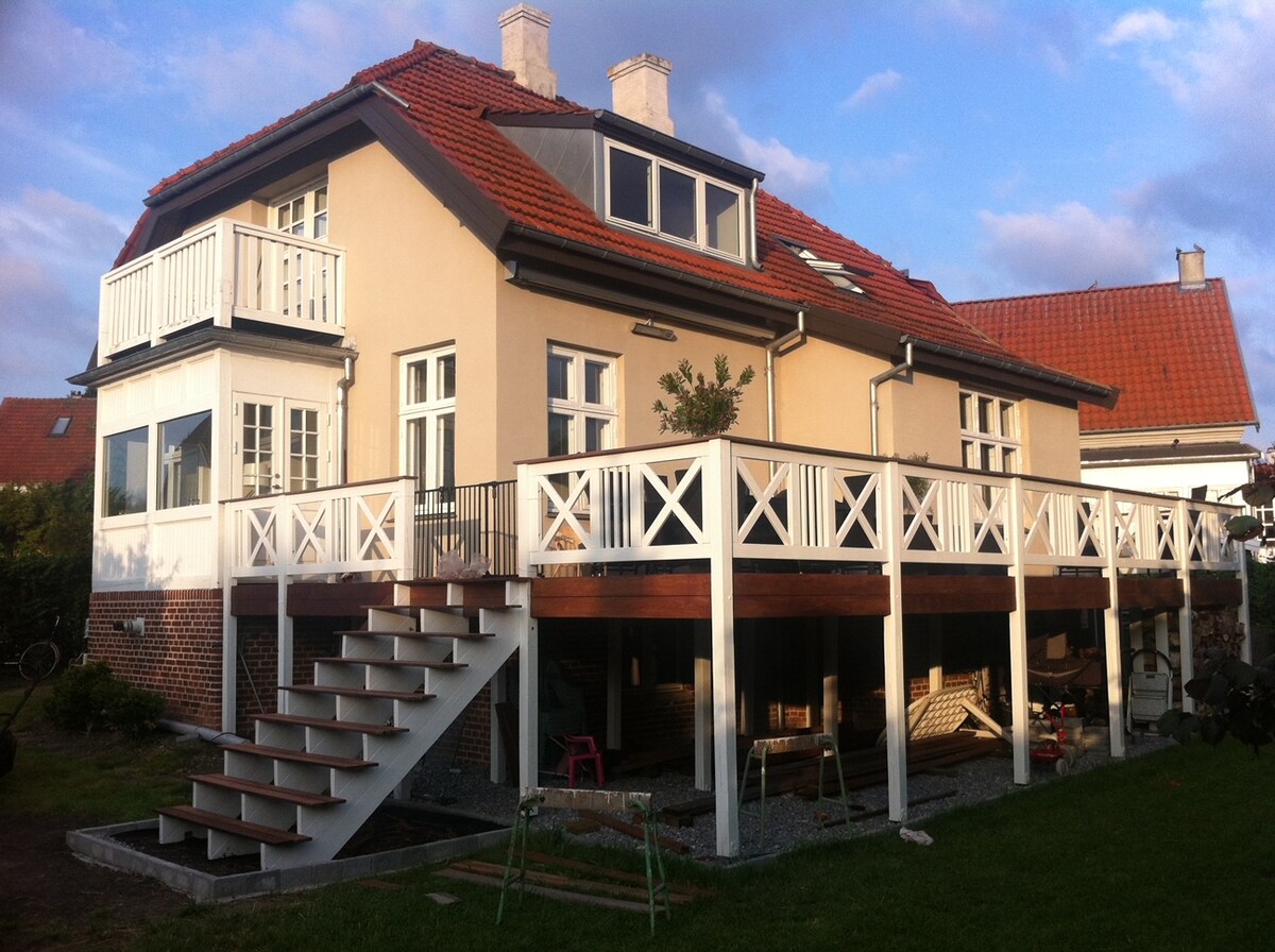The exterior of a charming two-story house is visible, featuring a large elevated deck with white railing and stairs leading down to the garden. Multiple windows allow natural light, and the roof is adorned with simple chimneys. A well-maintained lawn is present at ground level.