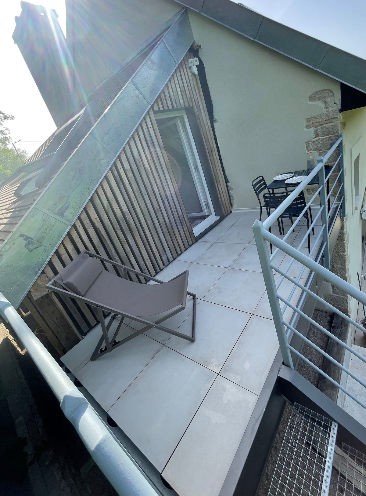 A private terrace is displayed, featuring a small round table and two chairs for outdoor seating. A folding lounge chair is positioned against the railing. Light tiles cover the floor, and the sloped roof adds architectural interest to the space.