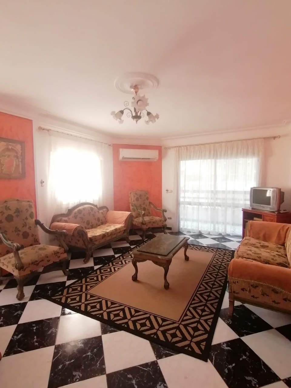 A bright living room features floral-patterned seating arranged around a central glass coffee table. The floor displays a bold black and white checkered pattern. Natural light fills the room through a large window, complemented by light sheer curtains. A small television is positioned on a wooden stand.