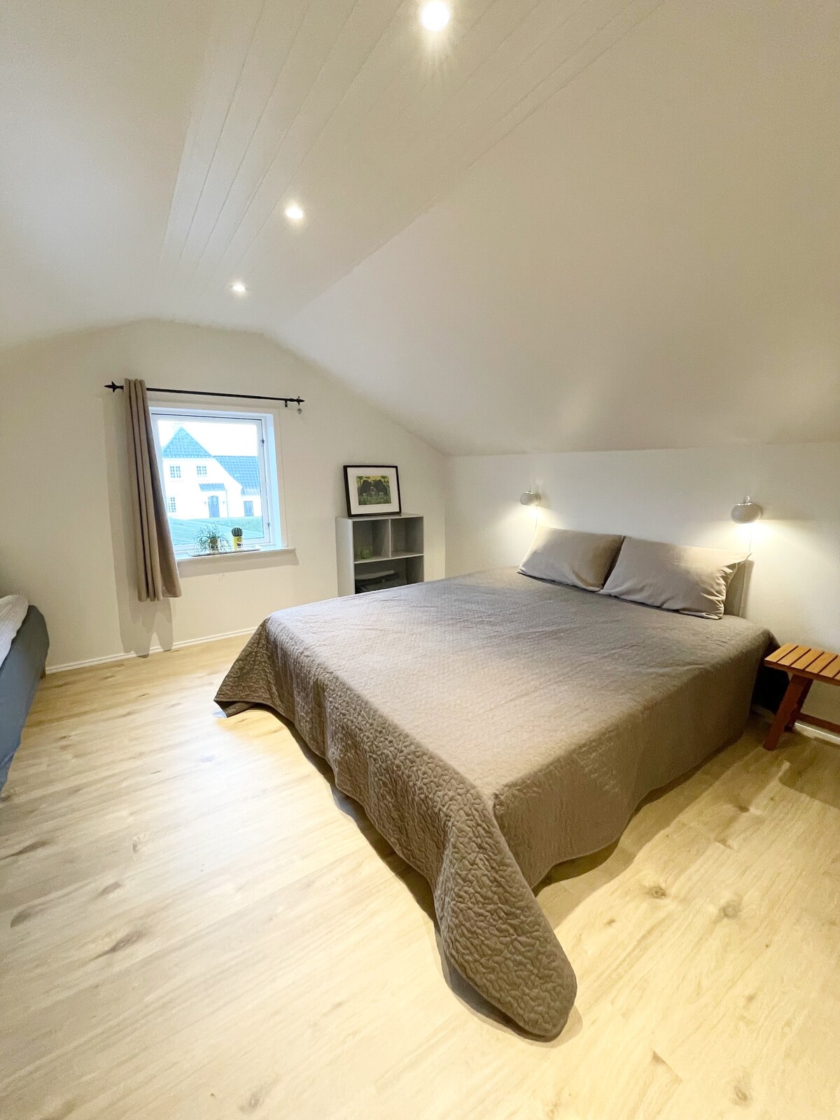 A well-lit room features a spacious bed with fresh linens and two wall-mounted lights for convenience. A window beside the bed provides natural light, and light-colored wooden flooring enhances the airy atmosphere. A small side table offers additional utility.