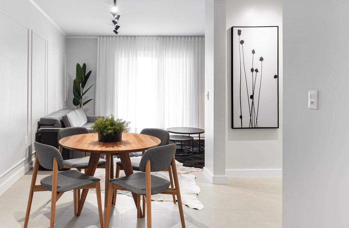 A modern dining area is presented with a round wooden table surrounded by four upholstered chairs. A potted plant is displayed at one side, while natural light filters through sheer curtains at the window, enhancing the neutral decor and minimalist wall art.