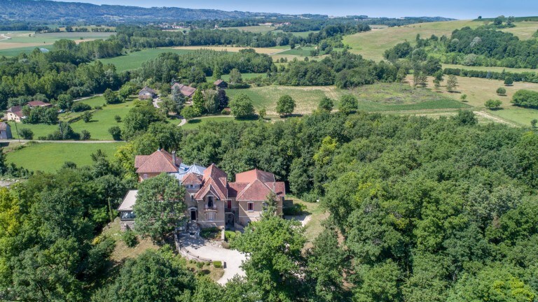 Aerial view of a spacious property surrounded by lush greenery and rolling hills. The structure features a variety of roof styles and is set among mature trees, offering a serene atmosphere. Open fields in the distance enhance the sense of seclusion.