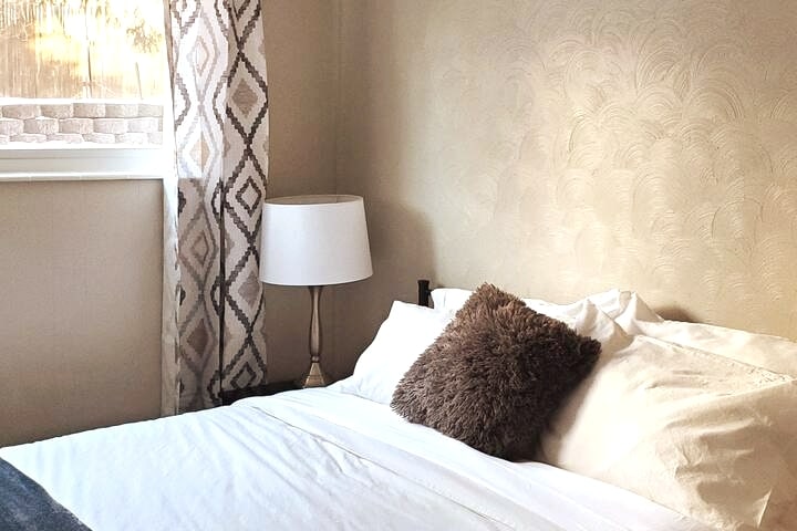 A spacious bed is presented with crisp white linens and a plush decorative pillow. Natural light filters through a nearby window, framed by patterned curtains. A bedside lamp complements the warm tones of the textured wall.