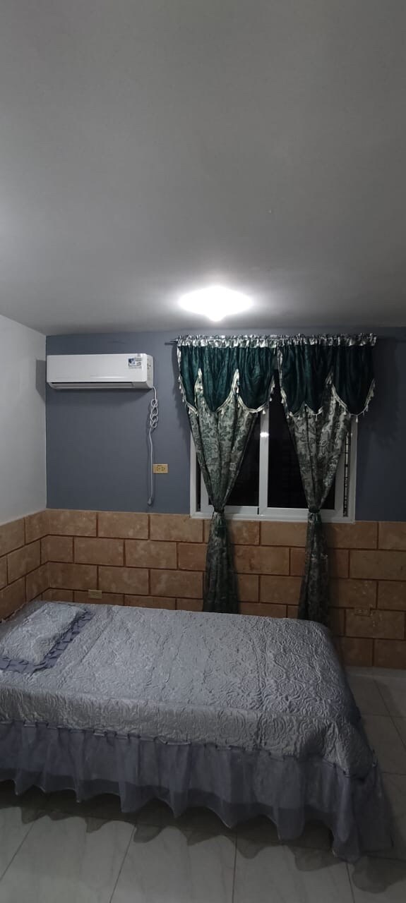 A modest bedroom features a spacious bed with a decorative cover, situated against a light wall with a textured stone base. Air conditioning is mounted on the wall, while elegantly draped green curtains frame a window, allowing soft light to enter the room.