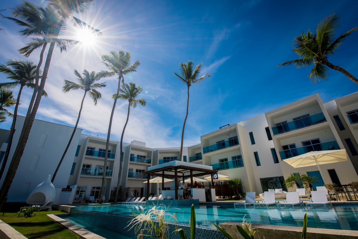 A clear blue sky is seen above, with the sun shining brightly over a modern resort building. Tall palm trees are positioned around a tranquil pool area, which features lounge chairs and a shaded seating structure, offering a comfortable space for relaxation.