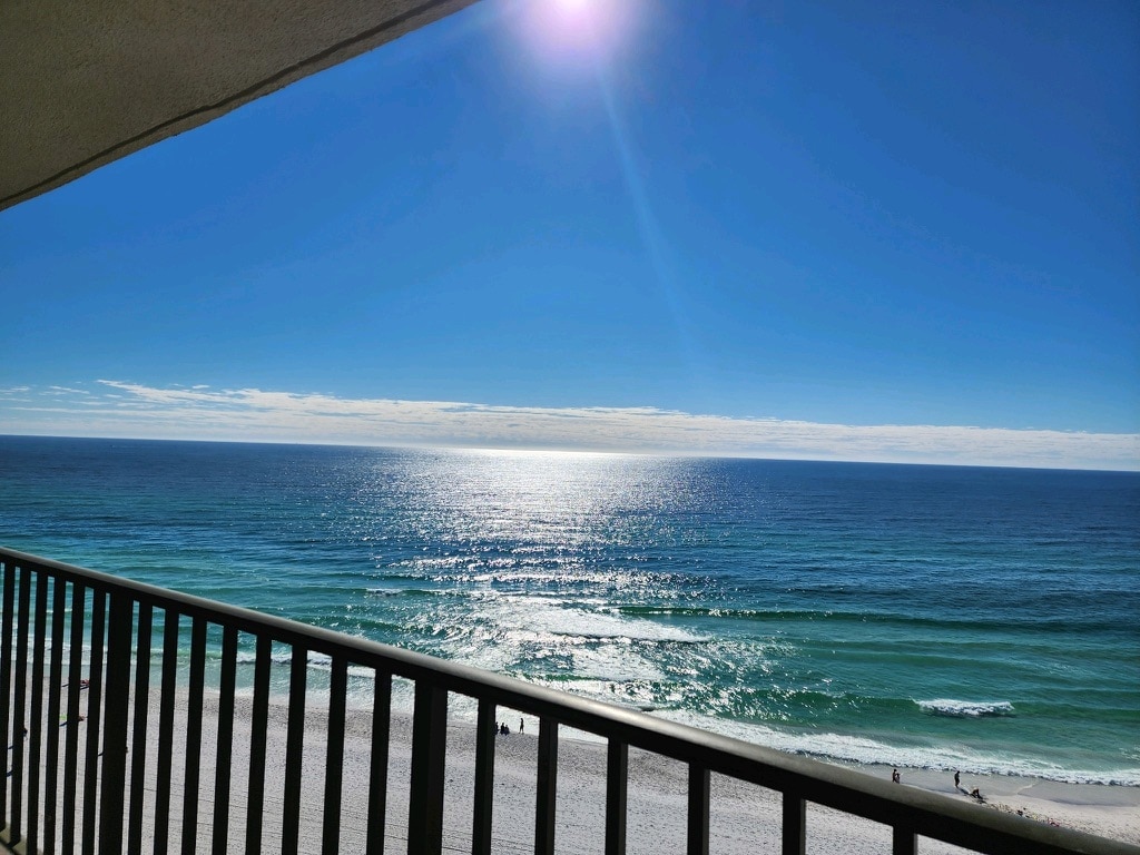The expansive view of the Gulf of Mexico highlights shimmering turquoise waters under a clear sky. Gentle waves lap against the sandy beach, while a distant shoreline can be seen. The balcony railing outlines the scene, framing the serene coastal landscape.