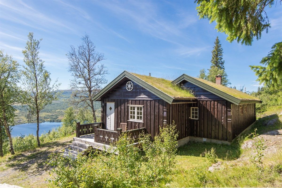 The charming wooden cabin features a grass-covered roof and traditional design elements. Surrounded by lush greenery, it offers views of the nearby lake and landscape. A small front porch with steps provides access to the entrance, adding to the inviting exterior.
