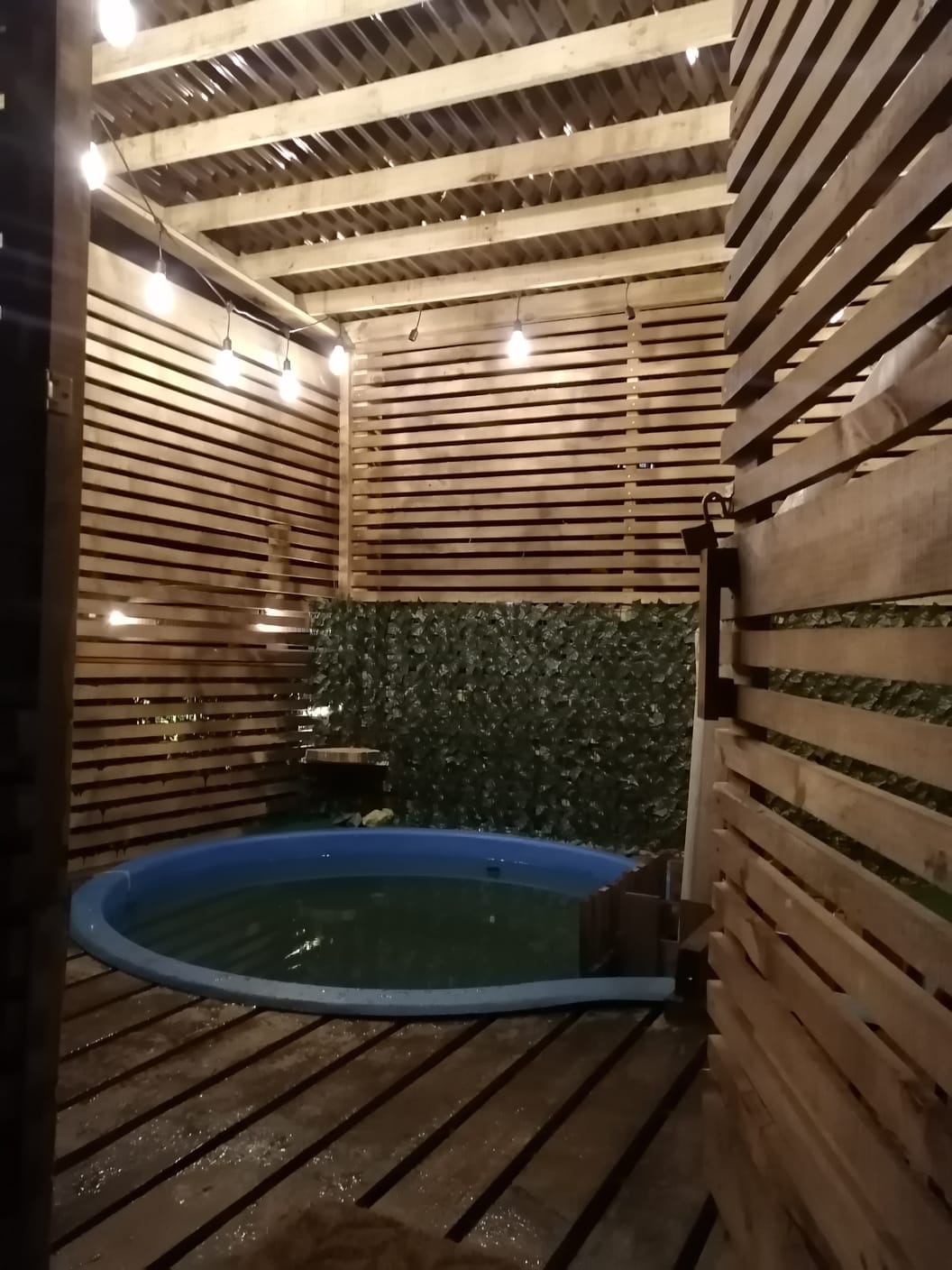 A private jacuzzi is surrounded by wooden slats and greenery, creating a secluded atmosphere. Soft lighting is provided by hanging bulbs overhead, illuminating the space gently. The blue interior of the jacuzzi reflects the light, enhancing the tranquil setting.