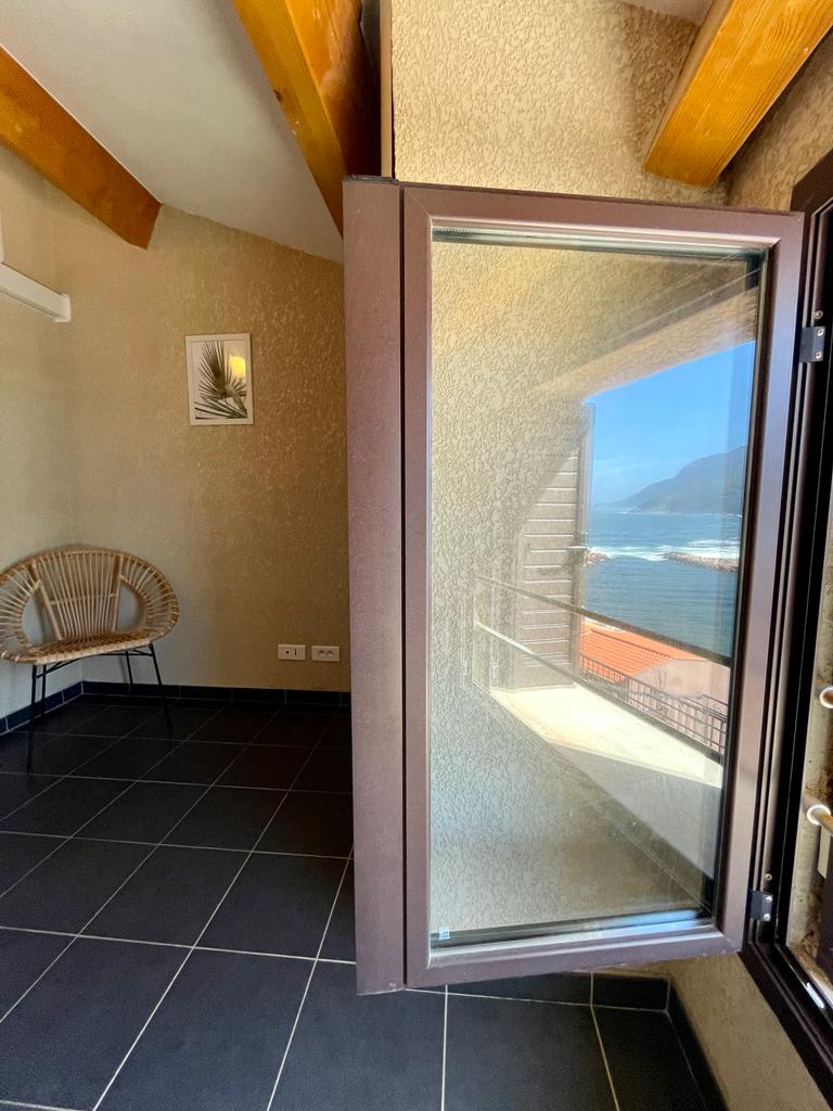 A corner of the room is highlighted by a large window, opening to a view of the sea and coastline. A chair with a woven design is positioned against the wall. Natural light fills the space, accentuating the room's textured walls and dark tiled floor.