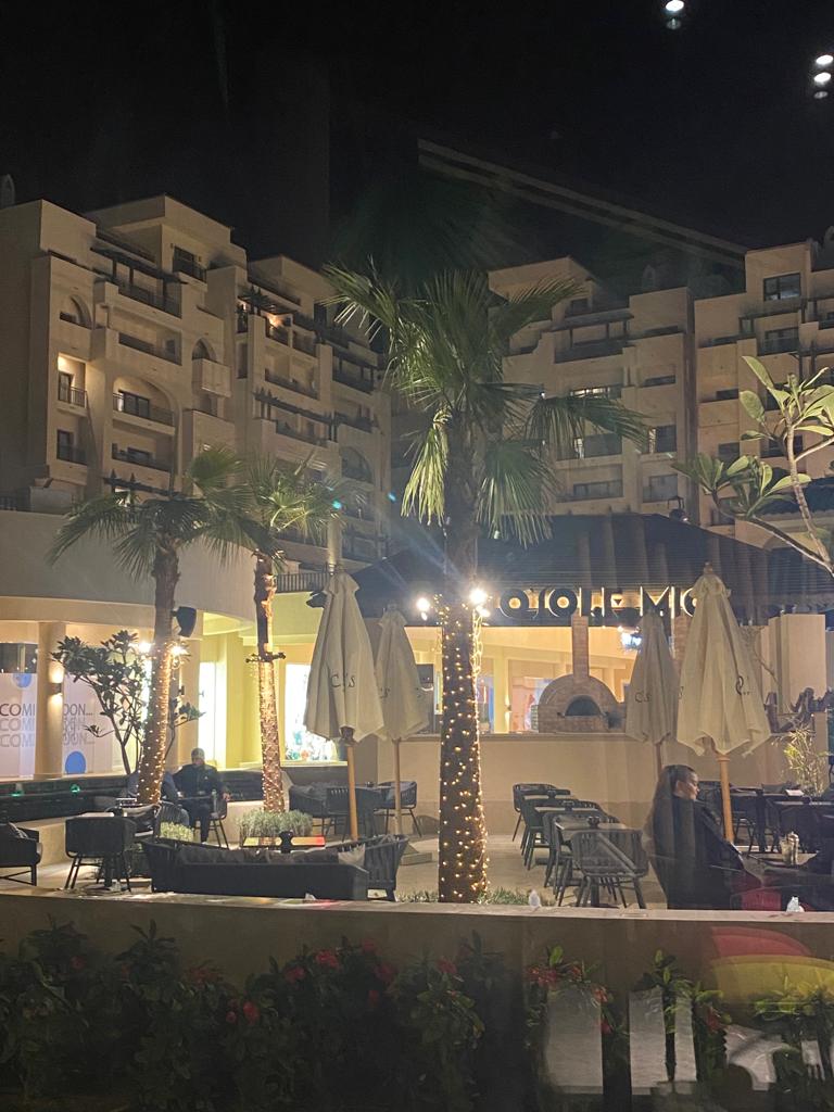 An outdoor dining area is depicted, featuring black wicker chairs and tables surrounded by well-maintained greenery and tropical palm trees. Soft lighting illuminates the space, creating a welcoming environment for guests. The modern architecture of the building is visible in the background.