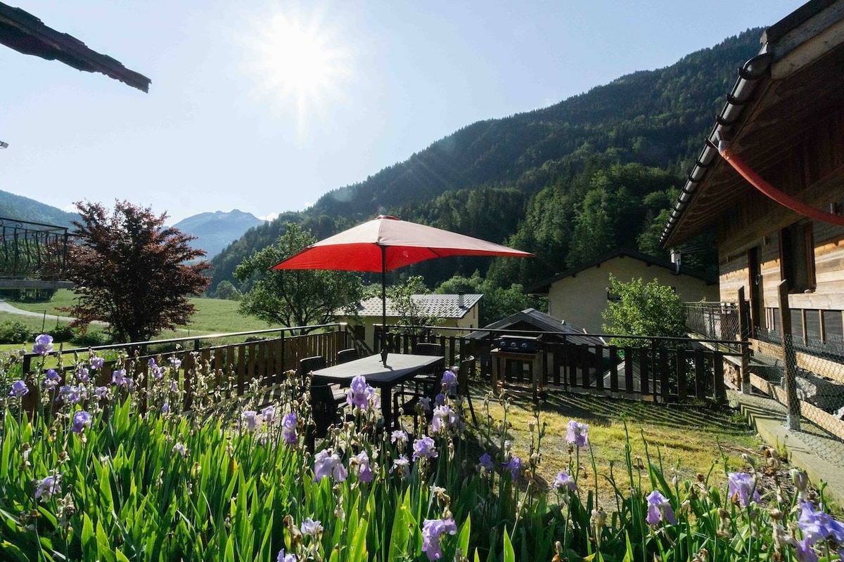 An outdoor terrace is presented with a table and chairs under a red umbrella. Surrounding greenery includes blooming flowers, while a scenic mountain view enhances the tranquil space. Sunlight filters through, creating a bright and inviting atmosphere.