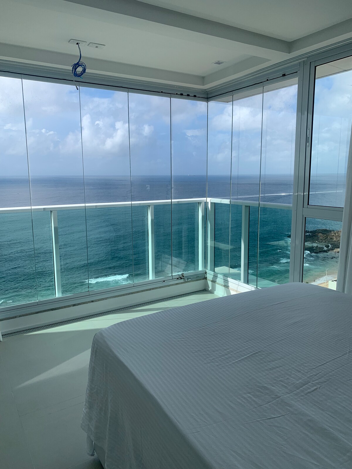 A bright bedroom features large glass walls offering an unobstructed view of the ocean. The bed is positioned to take advantage of natural light from the expansive windows, while the tranquil blue waters can be seen through the transparent barriers.