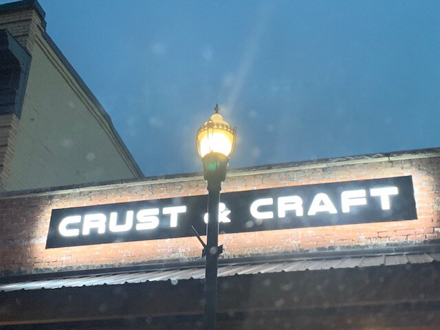 A restaurant sign is illuminated against a dusky blue sky, displaying the name 'Crust & Craft' in bold white letters. A street lamp casts a warm glow below, highlighting brick architecture and providing a sense of urban charm.