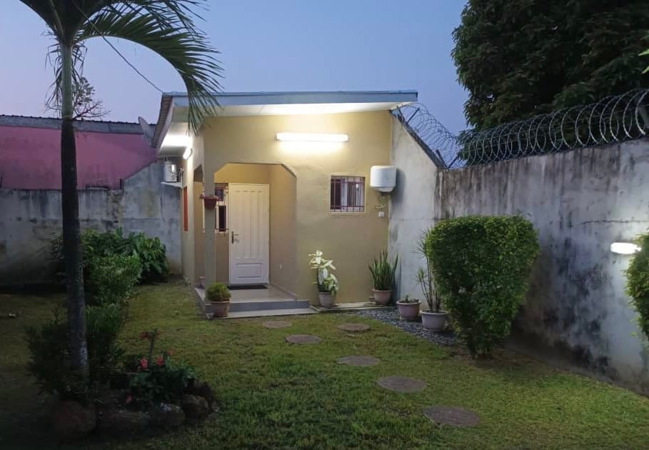 The exterior of the studio is framed by a well-kept garden, featuring a manicured lawn and potted plants. Soft lighting highlights the entrance, which is flanked by greenery, creating a welcoming ambiance as dusk sets in.