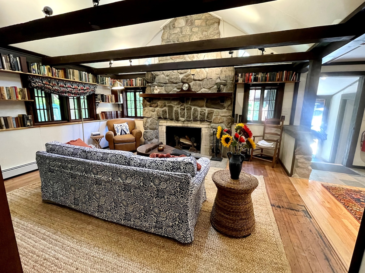 A cozy living area is highlighted by a stone fireplace and natural wood beams. A patterned sofa faces comfortable armchairs, with bookshelves filled with books along the walls. A round woven table holds a vase of fresh flowers, adding a touch of color to the space.