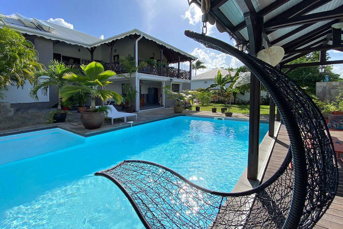 A spacious pool area is surrounded by tropical greenery, featuring a clear blue swimming pool. Comfortable loungers are positioned nearby, with a stylish, woven chair adding a touch of elegance. The villa is visible in the background, complemented by inviting landscaping.