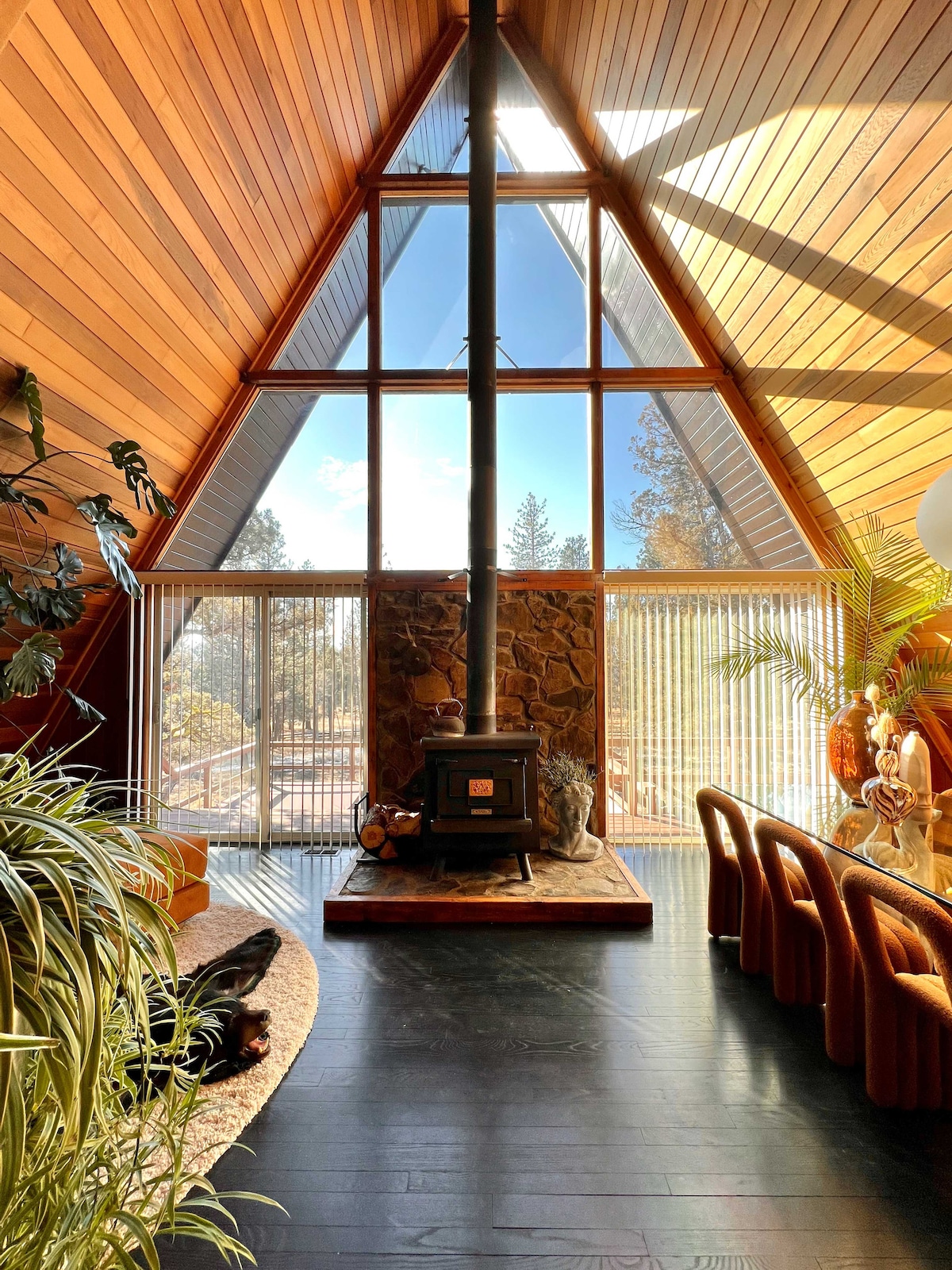 The interior of the A-frame cabin is highlighted by high wooden ceilings and large triangular windows, offering abundant natural light. A stone feature wall surrounds a wood stove, while a cozy seating area is arranged on a dark floor. Green plants add a touch of nature.