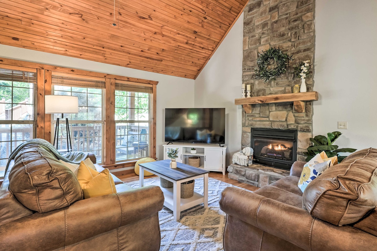 The living room features a warm wood ceiling and a stone fireplace, creating a cozy atmosphere. A large television is mounted on a white media console, with two comfortable sofas positioned around a central coffee table. Natural light enters through multiple windows, offering a view of the outdoors.