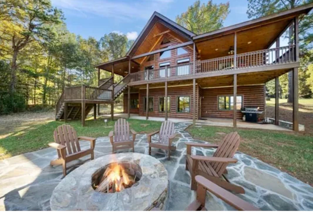 A spacious log cabin featuring an expansive deck and multiple large windows. An outdoor stone fire pit is surrounded by wooden chairs. A well-maintained lawn extends beyond the patio, complemented by mature trees in the background.