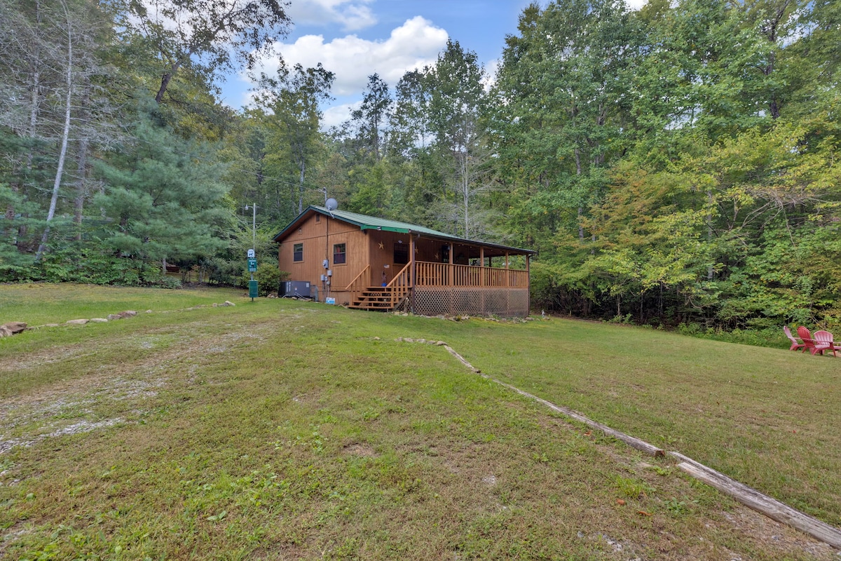A cabin is set amidst greenery, featuring a wooden deck with steps leading up to the entrance. Surrounding trees provide a natural backdrop, while an expanse of grassy area is visible in the foreground, offering a sense of seclusion.