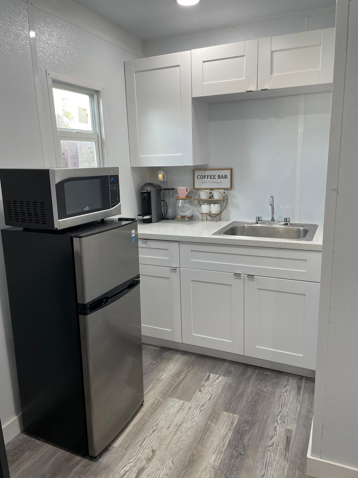 A compact kitchen space features sleek white cabinetry and a small countertop. A stainless steel refrigerator and microwave are noted, along with a coffee maker on the counter. A well-lit window provides natural light, enhancing the fresh and clean look of the area.