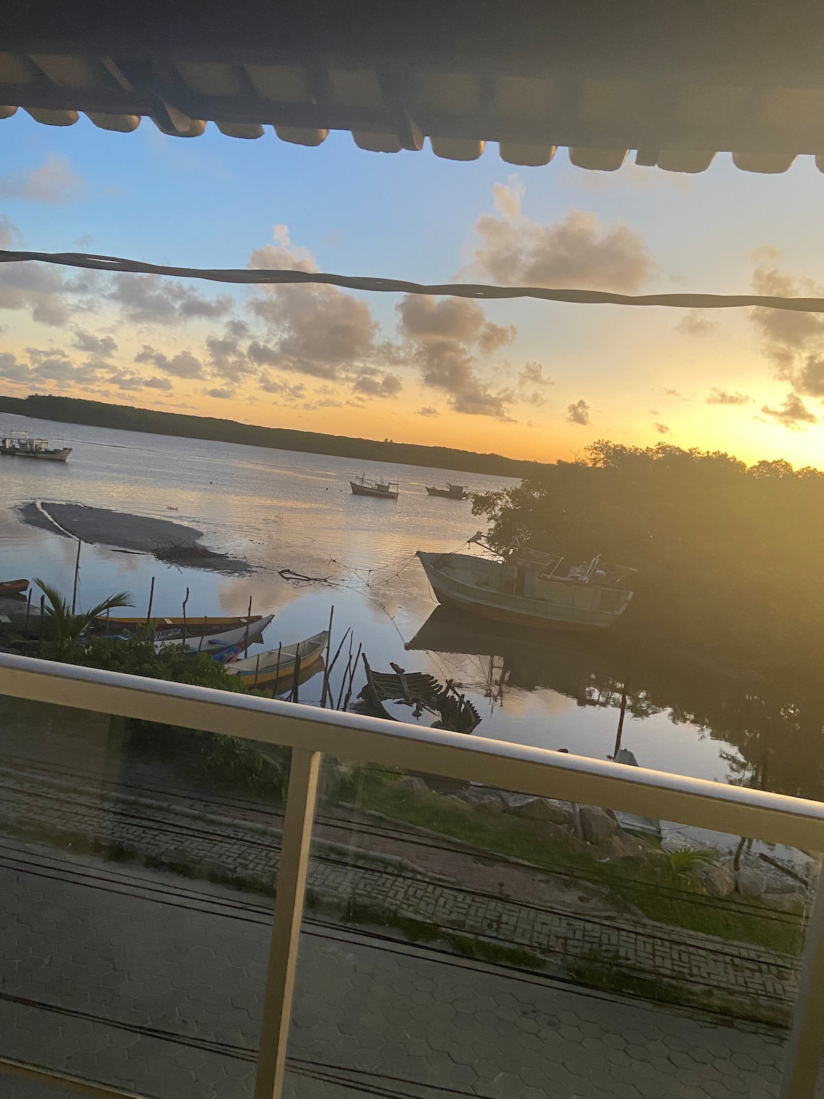 A serene view of the river at sunset is captured from a balcony. Boats are moored along the water's edge, with a soft glow illuminating the landscape. The sky is painted in hues of orange and blue, creating a calm atmosphere.