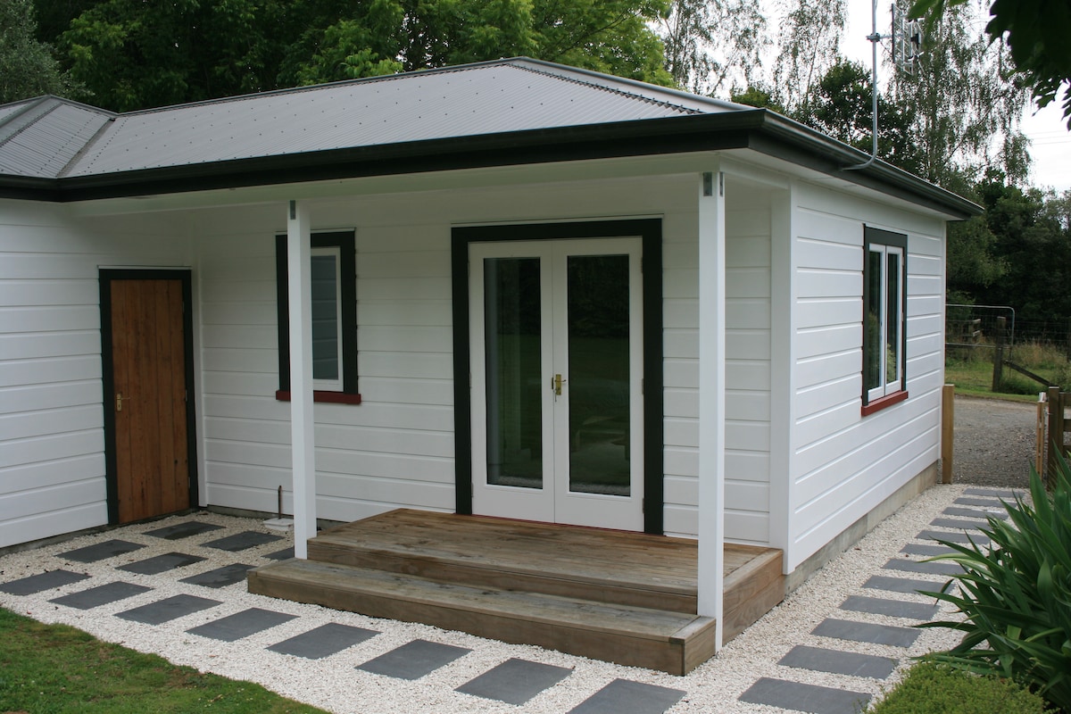The exterior of the cottage features a wooden porch with a simple, wide entrance and large glass doors framed by white walls. A pathway of rectangular stone tiles leads from the entrance, bordered by well-maintained gravel and greenery.