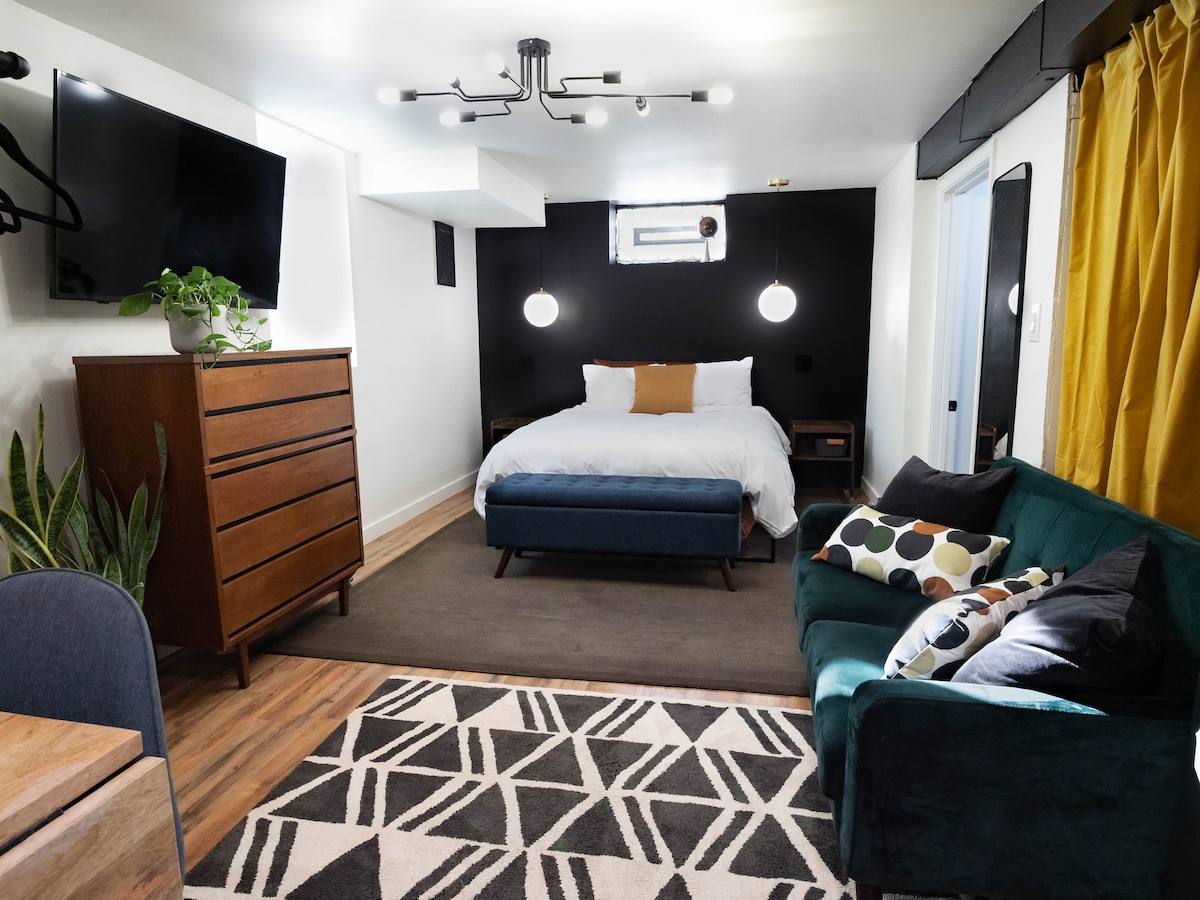 A stylish studio interior features a spacious bed with crisp white linens, complemented by a dark accent wall. A comfortable seating area with a green couch and patterned pillows is present. Additional furnishings include a wooden dresser, a table, and large windows providing natural light.