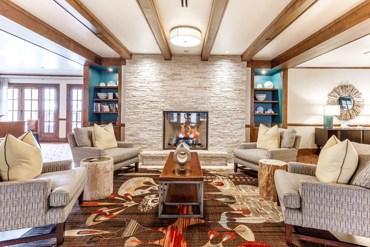 A cozy lounge area is highlighted by a modern gas fireplace, set within a stone feature wall. Two comfortable chairs with light-colored cushions are arranged around a wooden coffee table, complemented by a patterned area rug. Shelves displaying decorative items frame the fireplace.