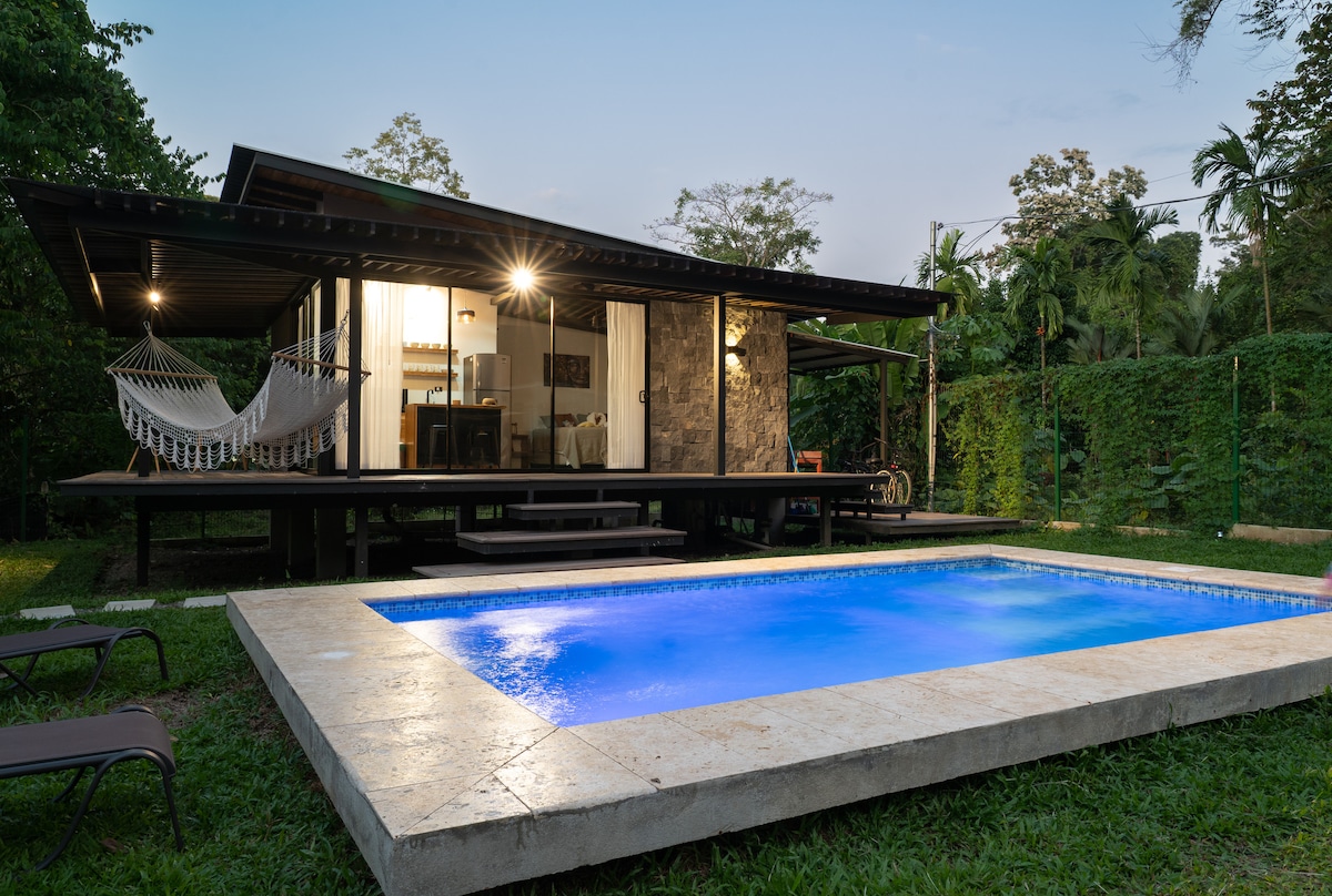 An eco-friendly cabin is positioned amidst a lush garden, featuring a swimming pool with illuminated blue waters. A hammock is seen strung between posts on the spacious deck, while lounge chairs are placed nearby. The cabin's modern design complements the tranquil surroundings.