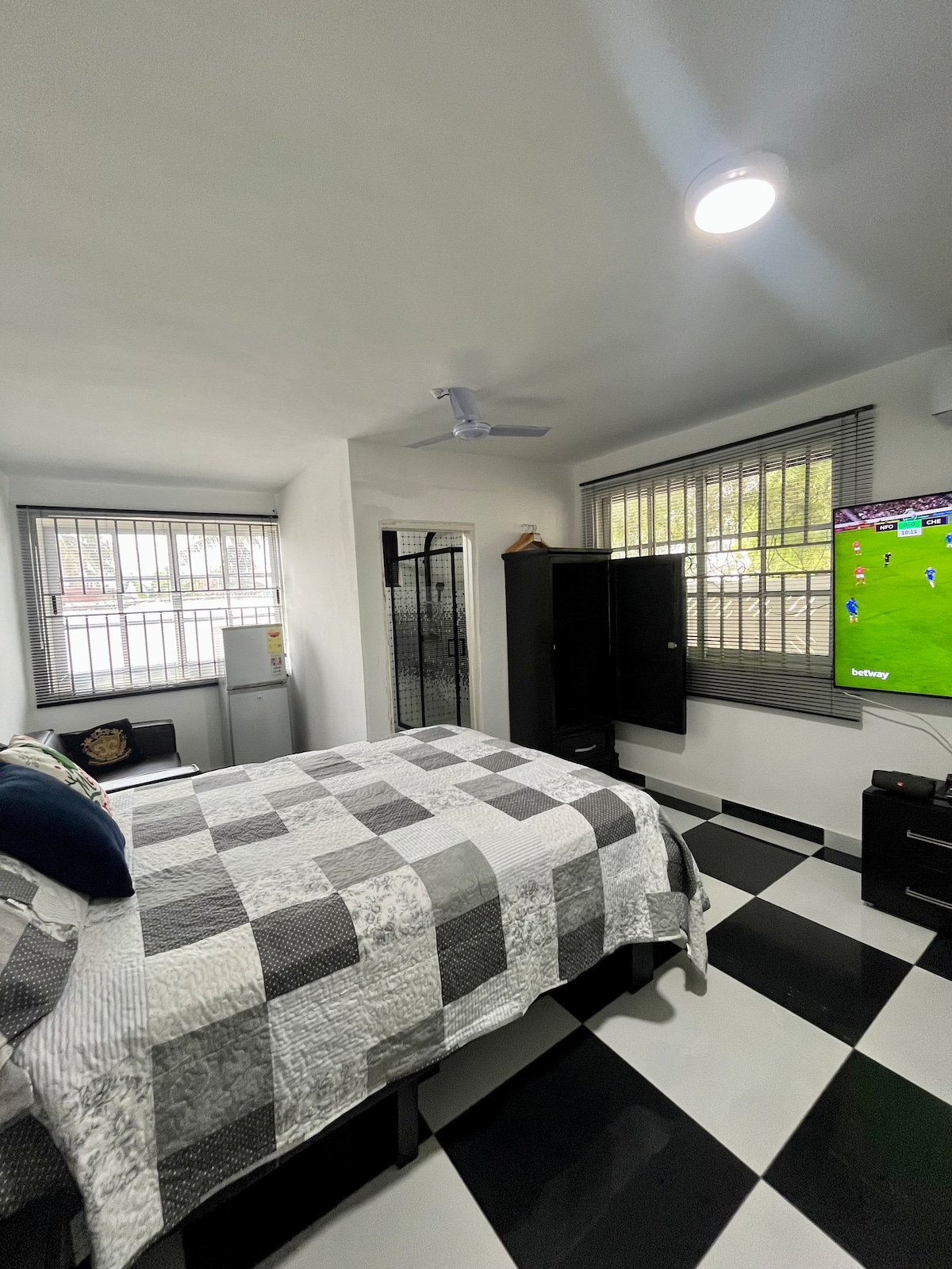 A well-lit bedroom features a spacious bed covered in a checkered quilt. A large wall-mounted TV displays a sports match. The room includes two windows with blinds, allowing natural light, and a closet stands against the wall, enhancing the overall functionality.