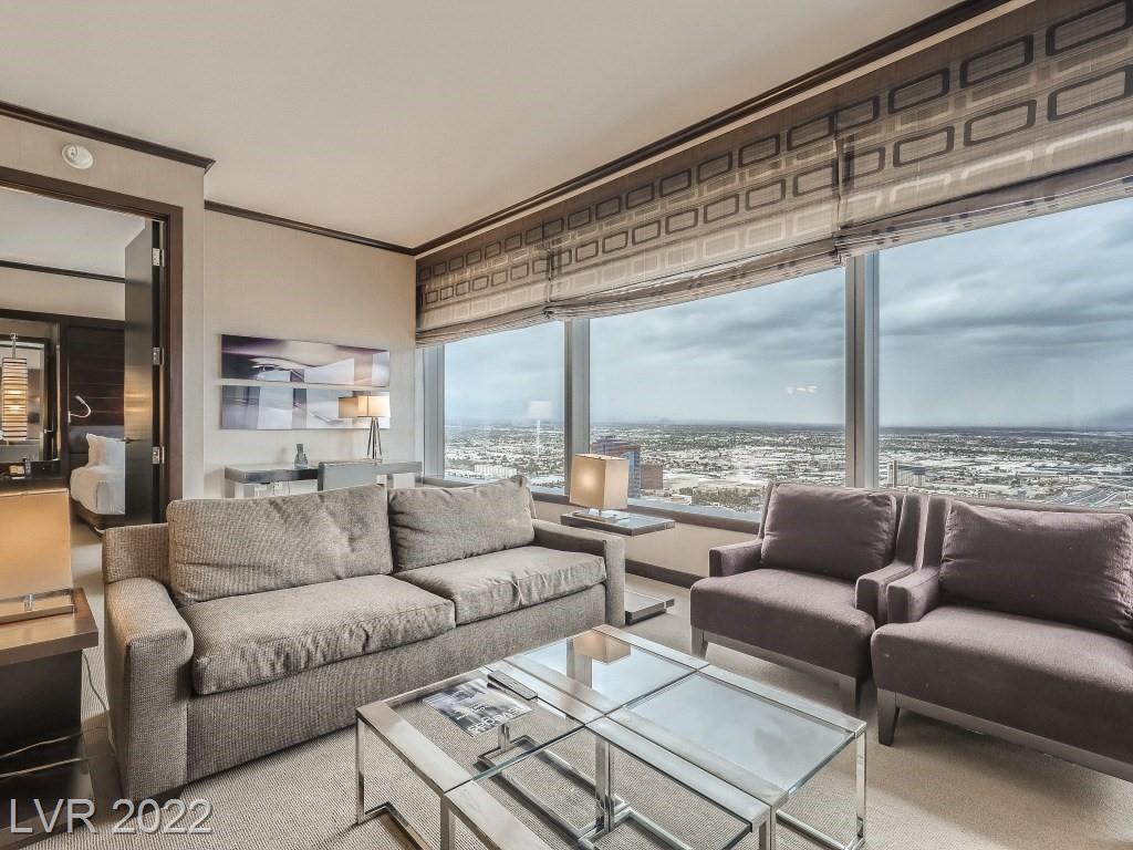 The living area features a comfortable gray sofa, accented by two armchairs in a muted tone. A glass coffee table sits in the center, with floor-to-ceiling windows offering expansive views of the surrounding cityscape.