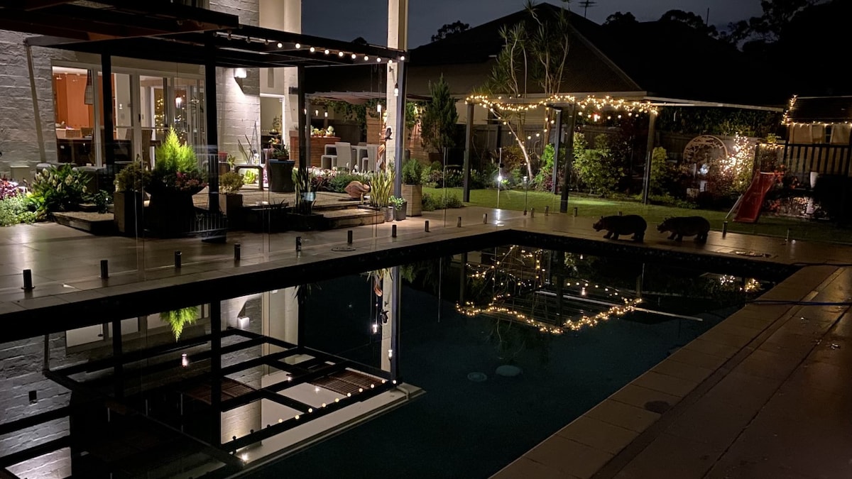 A calm evening scene highlights a private inground pool, surrounded by neatly landscaped gardens. Soft strings of lights illuminate the area, creating a serene atmosphere. The reflections in the water mirror the illuminated surroundings, including the house and greenery.