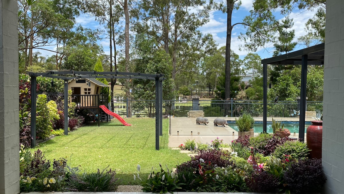 A view of the outdoor space features a manicured garden with vibrant plants and a grassy area. An enclosed play structure is visible, complete with a slide, while the inviting pool glimmers under the sun. Trees frame the serene landscape.