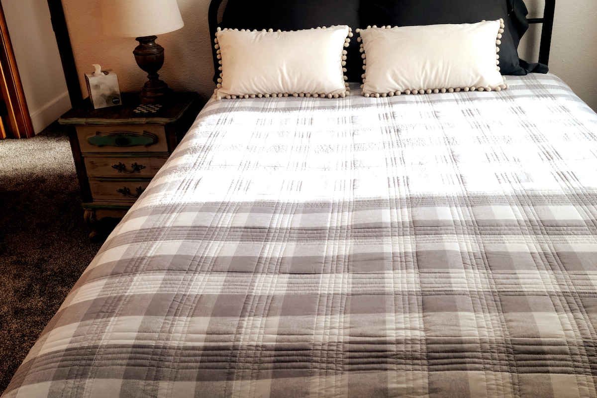 A neatly arranged bed features a plaid patterned comforter in shades of gray, with two decorative pillows resting on top. A bedside table holds a lamp and a small box, while soft light filters through the nearby window.