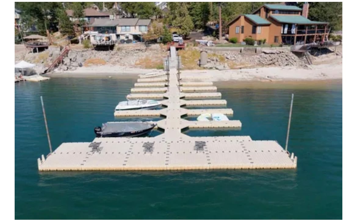 A boat dock extends into calm waters, lined with multiple docking spaces. Shoreline homes are visible in the background, surrounded by trees. The setting offers a serene view of Bass Lake, contributing to a peaceful lakeside atmosphere.