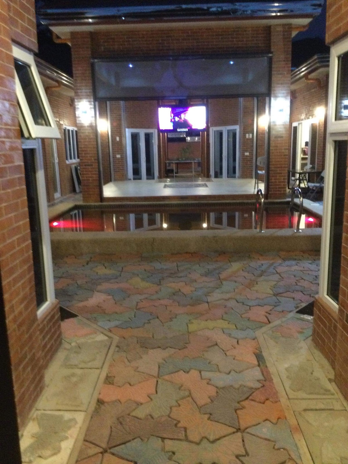 A vibrant courtyard area features a decorative mosaic floor in various colors. Large glass doors are visible, leading to a spacious interior with a television against a brick wall. Soft lighting enhances the ambiance, creating a relaxing environment.