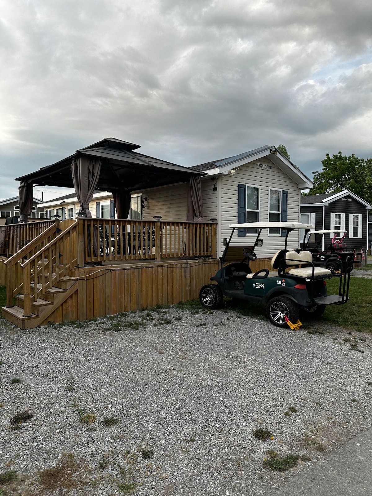 An outdoor space is highlighted by a wooden deck featuring a shaded area with draped curtains. A golf cart is parked nearby on gravel, providing easy access to the unit. The exterior of the unit is characterized by light-colored siding and nearby residential structures.