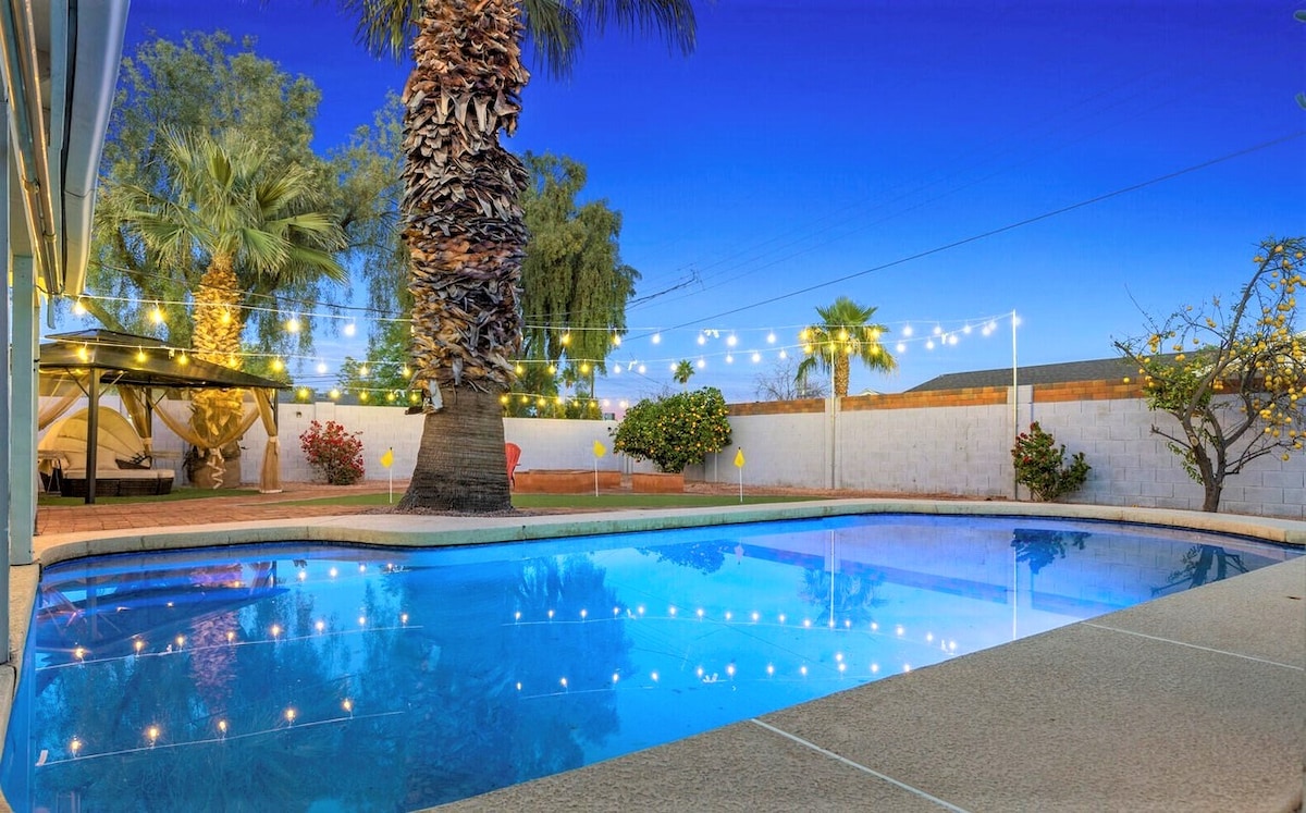 The backyard features a private pebble-tec pool under a clear twilight sky, surrounded by vibrant palm and fruit trees. String lights illuminate the space, creating a serene atmosphere. A shaded cabana is visible to one side, offering a cozy retreat within the lush garden.