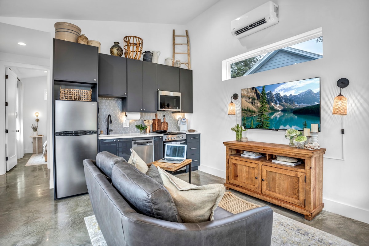 An open living space features a modern kitchen with stainless steel appliances and sleek cabinetry. A comfortable sofa is arranged in front of a wooden media console, which holds a large flat-screen TV. Natural light floods through a window, brightening the minimalist decor.