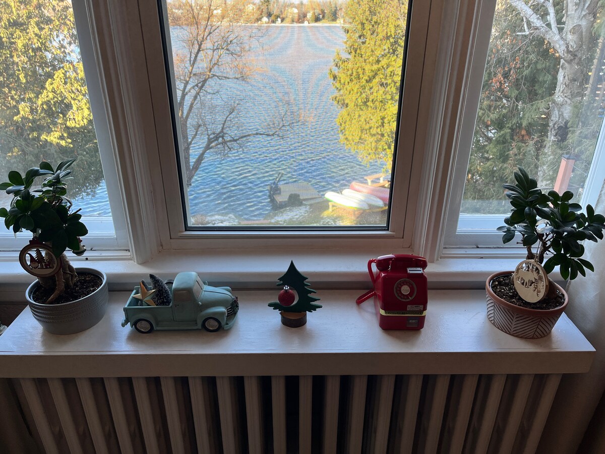 A bright window view features three decorative planters and a vintage toy truck. A small evergreen tree ornament and a red toy phone are placed on the windowsill, with a serene lake visible in the background, showcasing the natural setting surrounding the house.