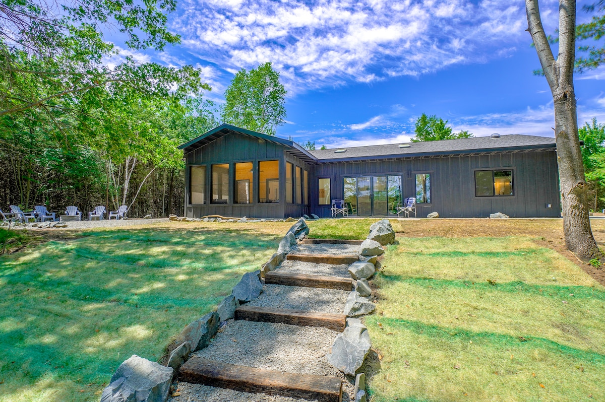 A modern lake house is framed by lush greenery and a well-maintained lawn. Steps made of gravel lead toward the building, which features large windows for natural light. Outdoor seating areas are visible, inviting relaxation in the scenic surroundings.