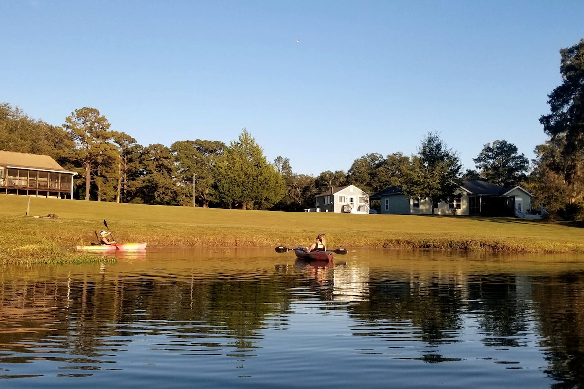 Two kayakers are gently paddling on a calm pond, surrounded by lush green grass and trees. The perspective captures a serene scene with a cottage and another building visible in the background under a clear blue sky.