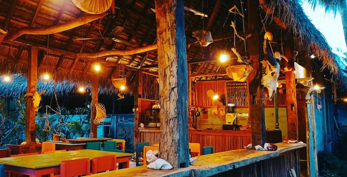 An open-air dining area is presented with a thatched roof supported by wooden beams. Colorful chairs surround a long wooden table. Decorative fish are suspended from the ceiling, and warm lights create a welcoming ambiance in the space.