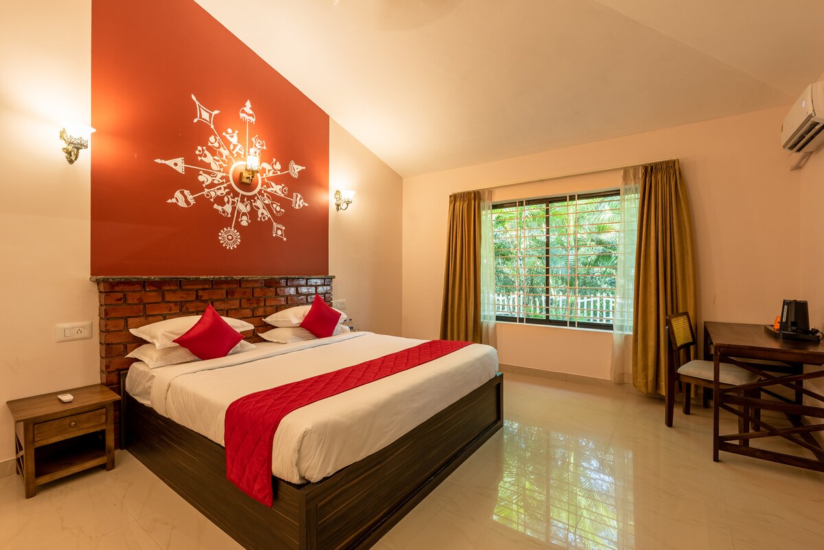A spacious room features a large bed adorned with red and white linens, complemented by decorative pillows. A brick accent wall adds character, while natural light floods in through a large window, offering a view of lush greenery outside. A wooden desk and chair provide a functional workspace.