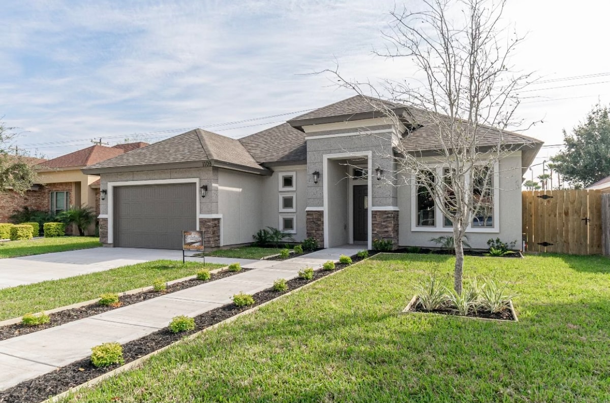 A modern single-story home is presented with a well-manicured lawn. The entrance features a covered porch with decorative stone accents. A two-car garage is visible to the left, complemented by a clean driveway and landscaped flower beds along the walkway.