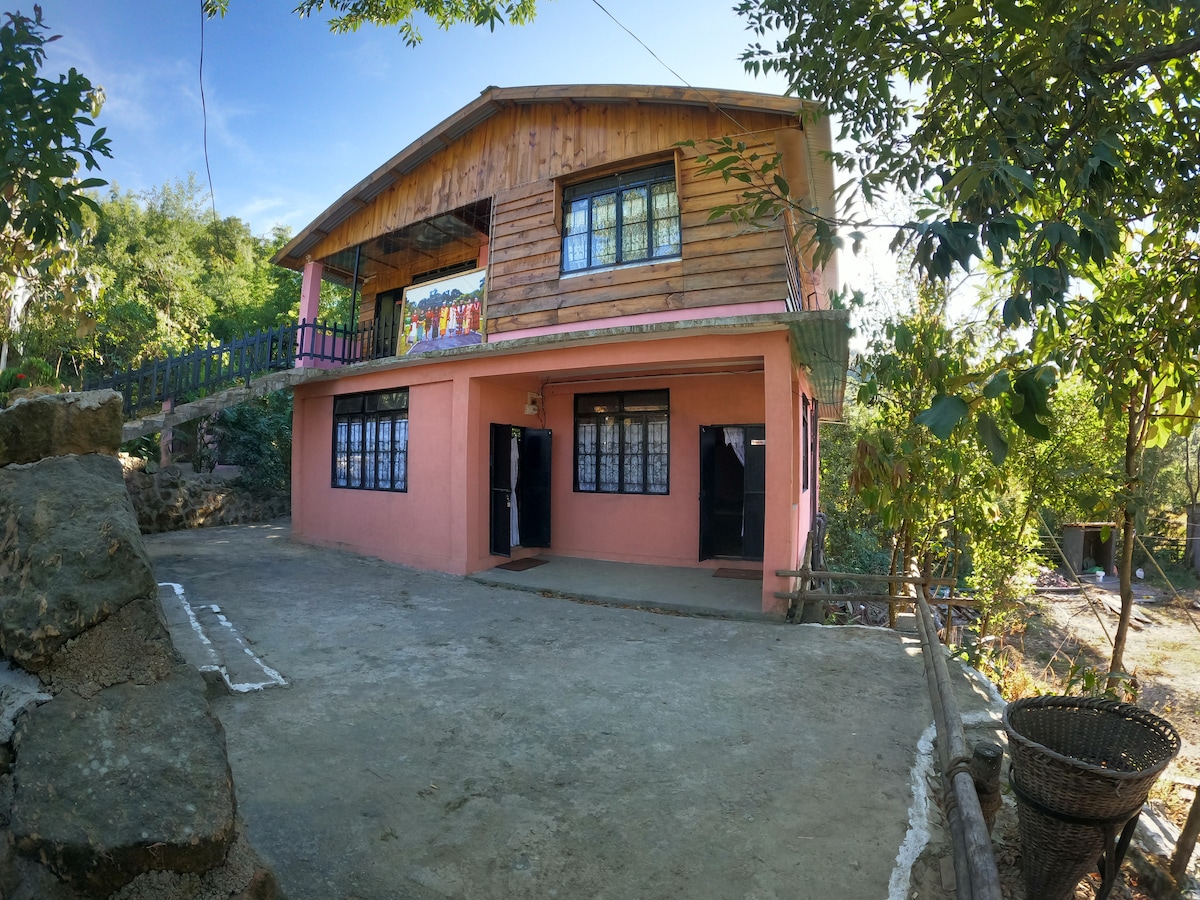 The guesthouse features a two-story wooden and concrete structure, with a welcoming pink façade. Large windows with delicate lace curtains provide natural light to the interiors. Surrounding greenery adds to the serene environment, with a stone path leading to the entrance.